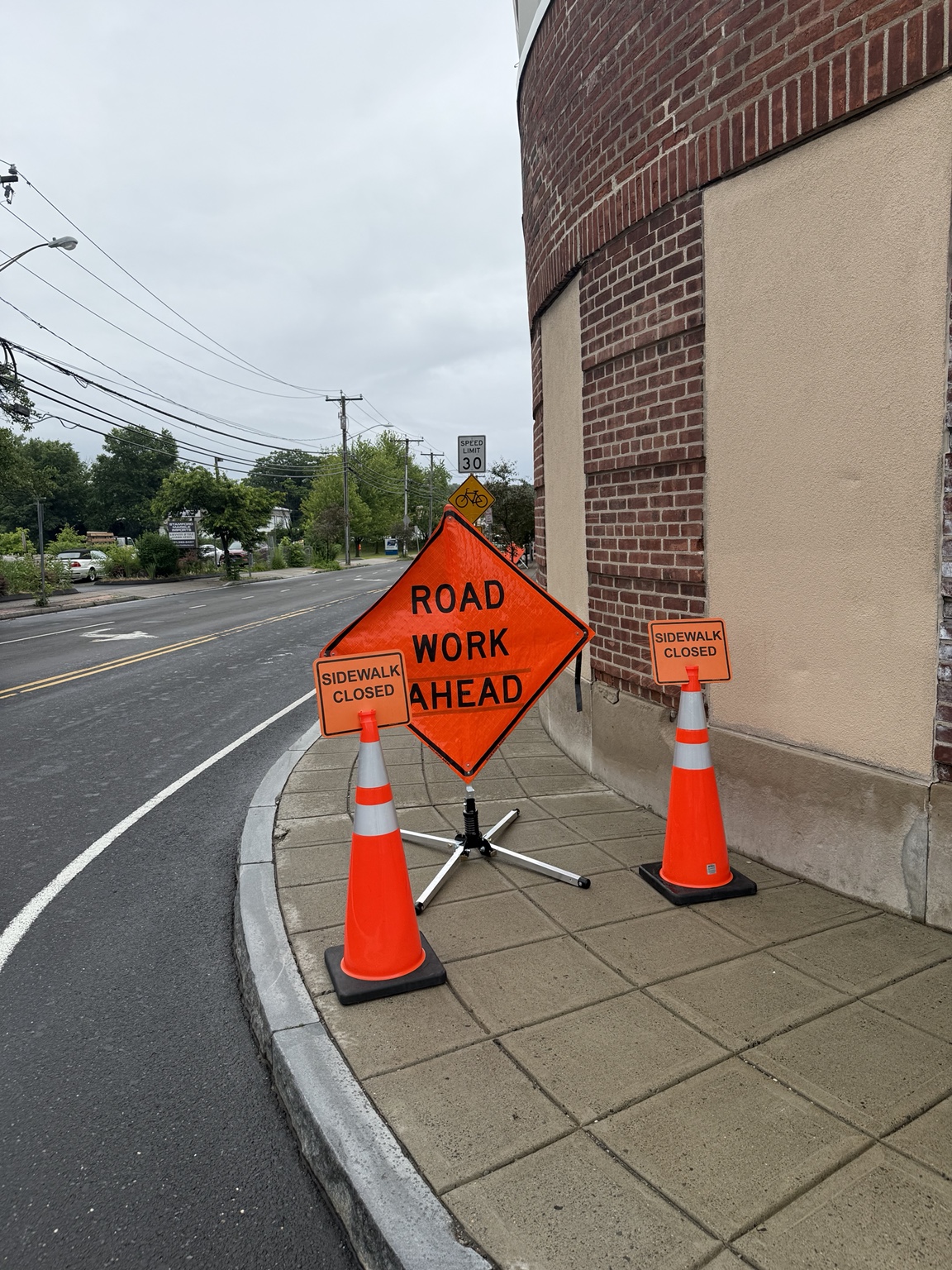 Road work and sidewalk closed signs with cones beside a brick building.